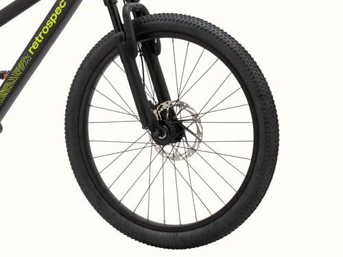 Close-up of the Koda Plus 24" Kids' Matte Black Mamba Bicycle's front wheel with disc brake and part of the frame visible displaying "retrospec" in neon green.