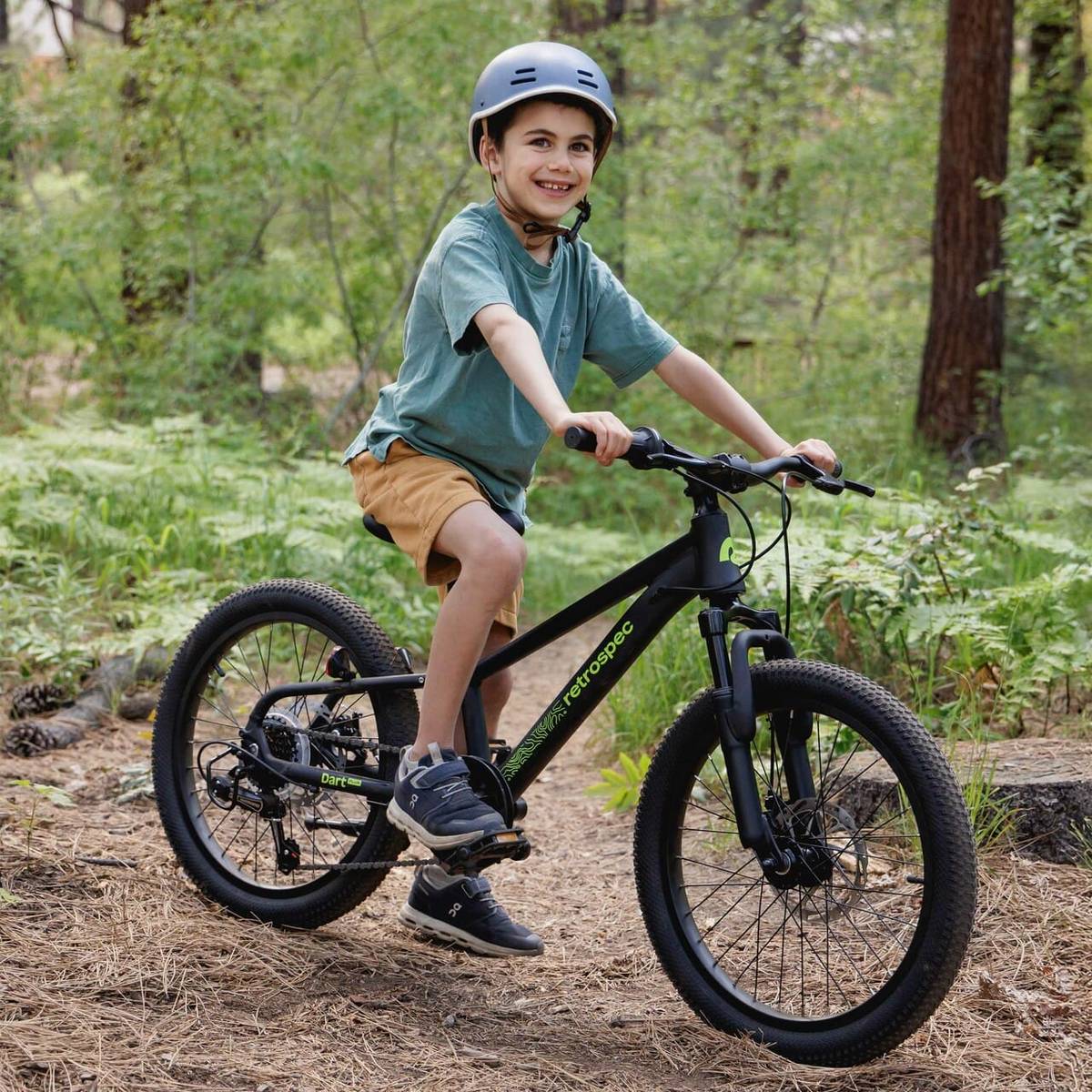 A child rides a black Dart Plus 20" bike in a lush, green forest setting, wearing a helmet and casual clothing.
