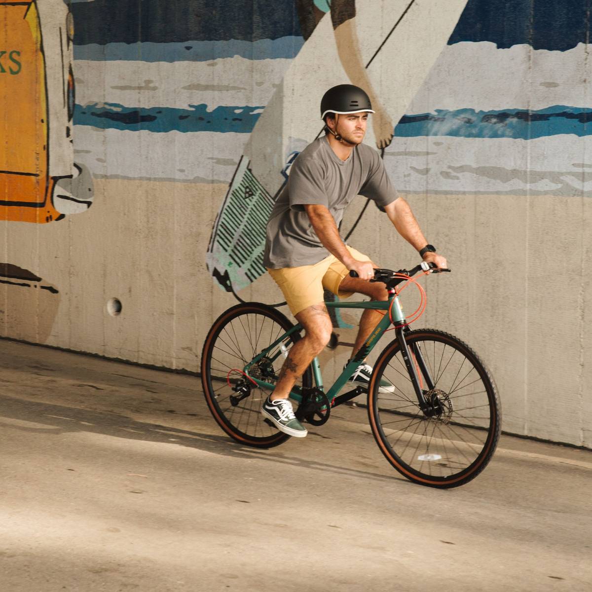A man rides a green Grateful Dead Amok Gravel Adventure Bike - 8 Speed on a sunny path with a surf ocean mural in the background, wearing a grey shirt, yellow shorts, and a black helmet, with a visible tattoo on his leg.