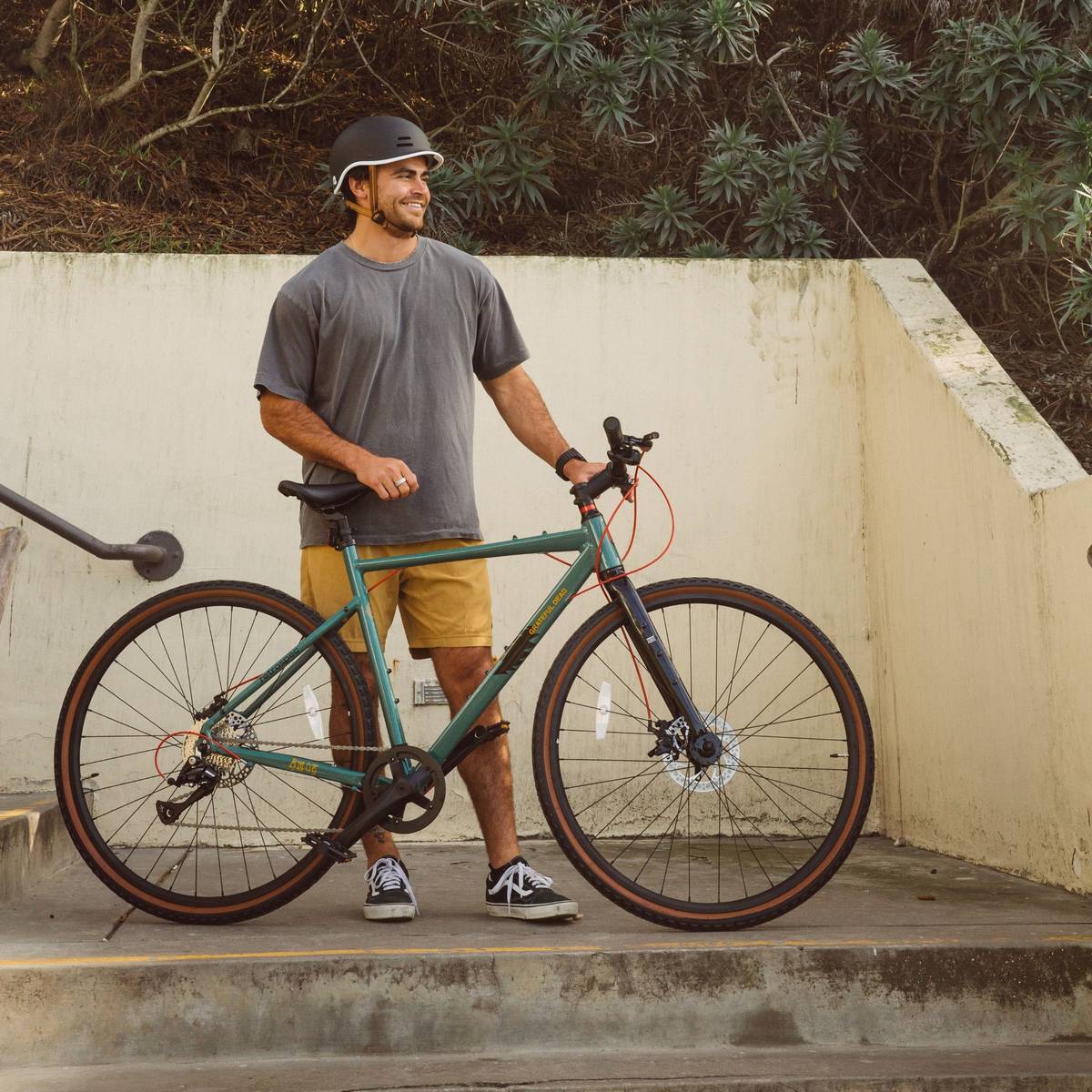 A person in a gray t-shirt and yellow shorts stands near stairs with the Grateful Dead Amok Gravel Adventure Bike 8-Speed.
