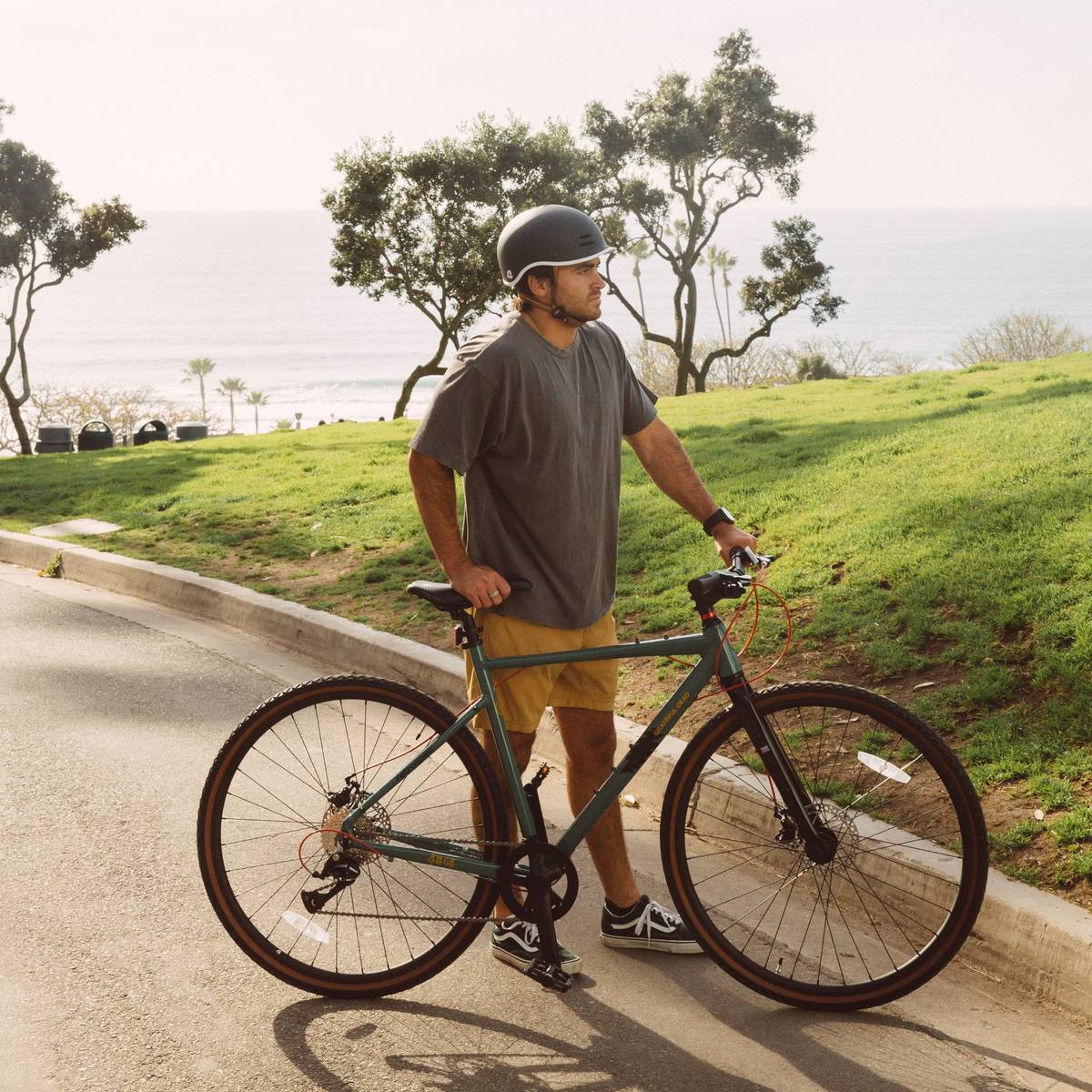 A person in a gray t-shirt and yellow shorts stands  with the Grateful Dead Amok Gravel Adventure Bike 8-Speed on a paved path with lush green grass in the background.