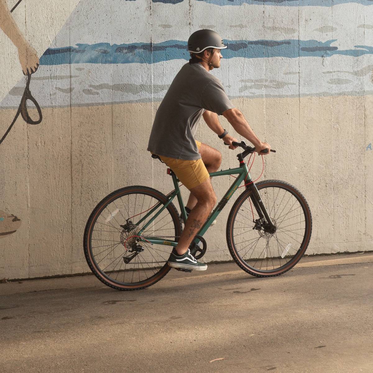 A man rides a green Grateful Dead Amok Gravel Adventure Bike - 8 Speed on a sunny path with a surf ocean mural in the background, wearing a grey shirt, yellow shorts, and a black helmet, with a visible tattoo on his leg.