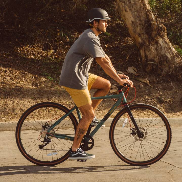 A man rides a green Grateful Dead Amok Gravel Adventure Bike - 8 Speed on a sunny path, wearing a grey shirt, yellow shorts, and a black helmet, with a visible tattoo on his leg.