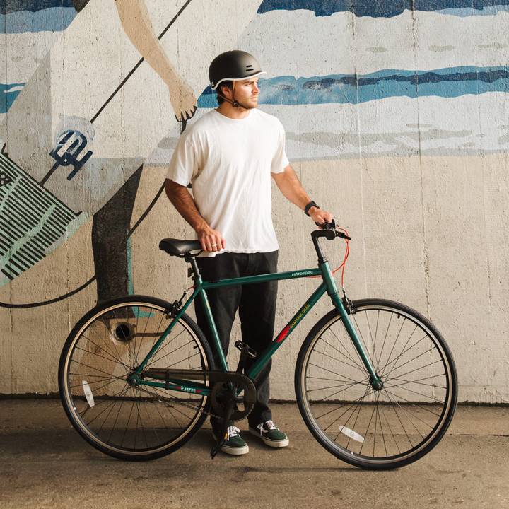 A person in a white t-shirt, black pants, and a helmet stands with a green Grateful Dead Harper Fixie Bike - Single Speed on a path, with a surf and ocean mural in the background.