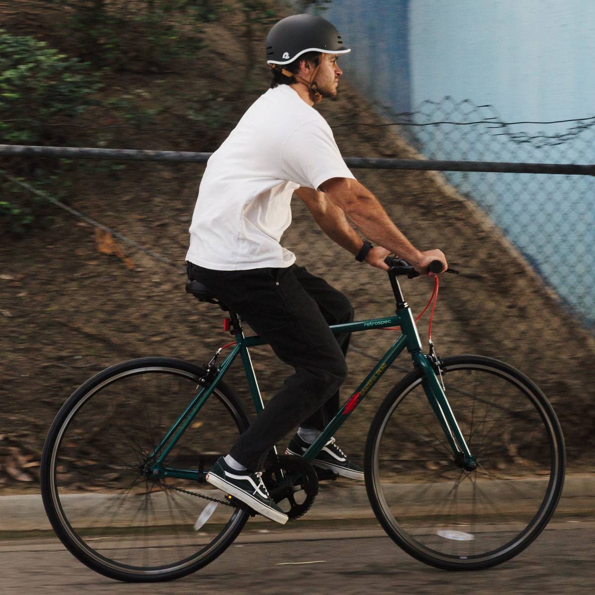A person in a white t-shirt and helmet rides a green Grateful Dead Harper Fixie Bike - Single Speed on a path, surrounded by trees and a blue wall in the background.