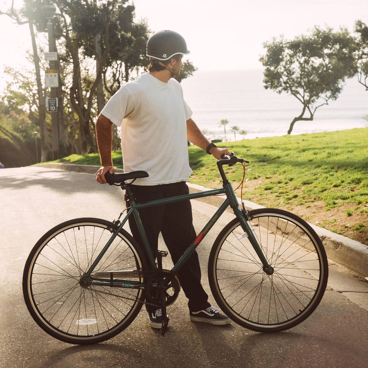 A person in a white t-shirt, black pants, and a helmet stands with a green Grateful Dead Harper Fixie Bike - Single Speed on a path, with a lush green grass and trees in the background.
