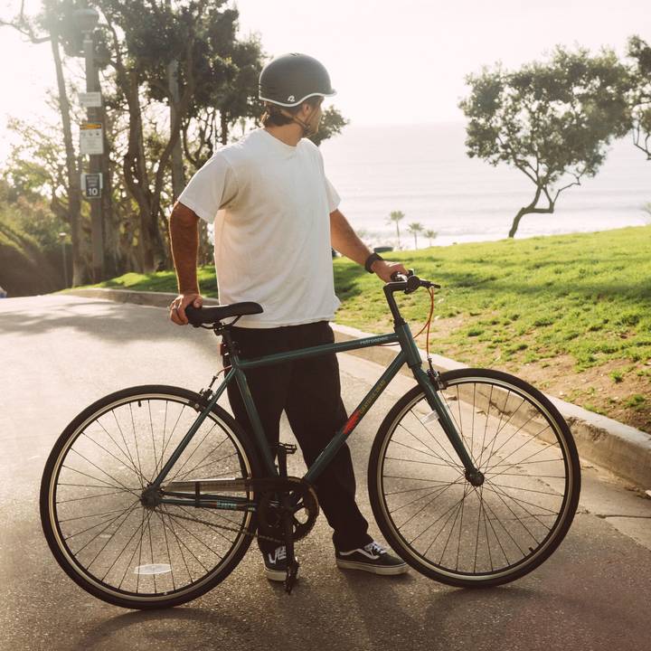 A person in a white t-shirt, black pants, and a helmet stands with a green Grateful Dead Harper Fixie Bike - Single Speed on a path, with a lush green grass and trees in the background.