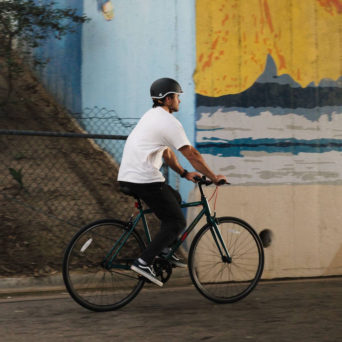 A person in a white t-shirt and helmet rides a green Grateful Dead Harper Fixie Bike - Single Speed on a path, surrounded by trees and a blue and yellow mural in the background.
