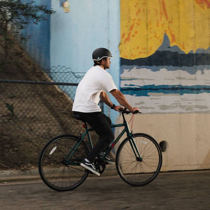 A person in a white t-shirt and helmet rides a green Grateful Dead Harper Fixie Bike - Single Speed on a path, surrounded by trees and a blue and yellow mural in the background.