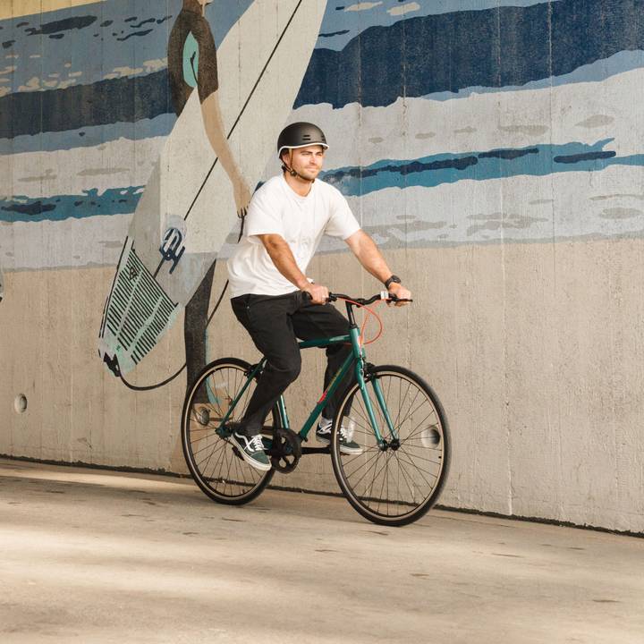 A person in a white t-shirt and helmet rides a green Grateful Dead Harper Fixie Bike - Single Speed on a path, with a surf and ocean mural in the background.