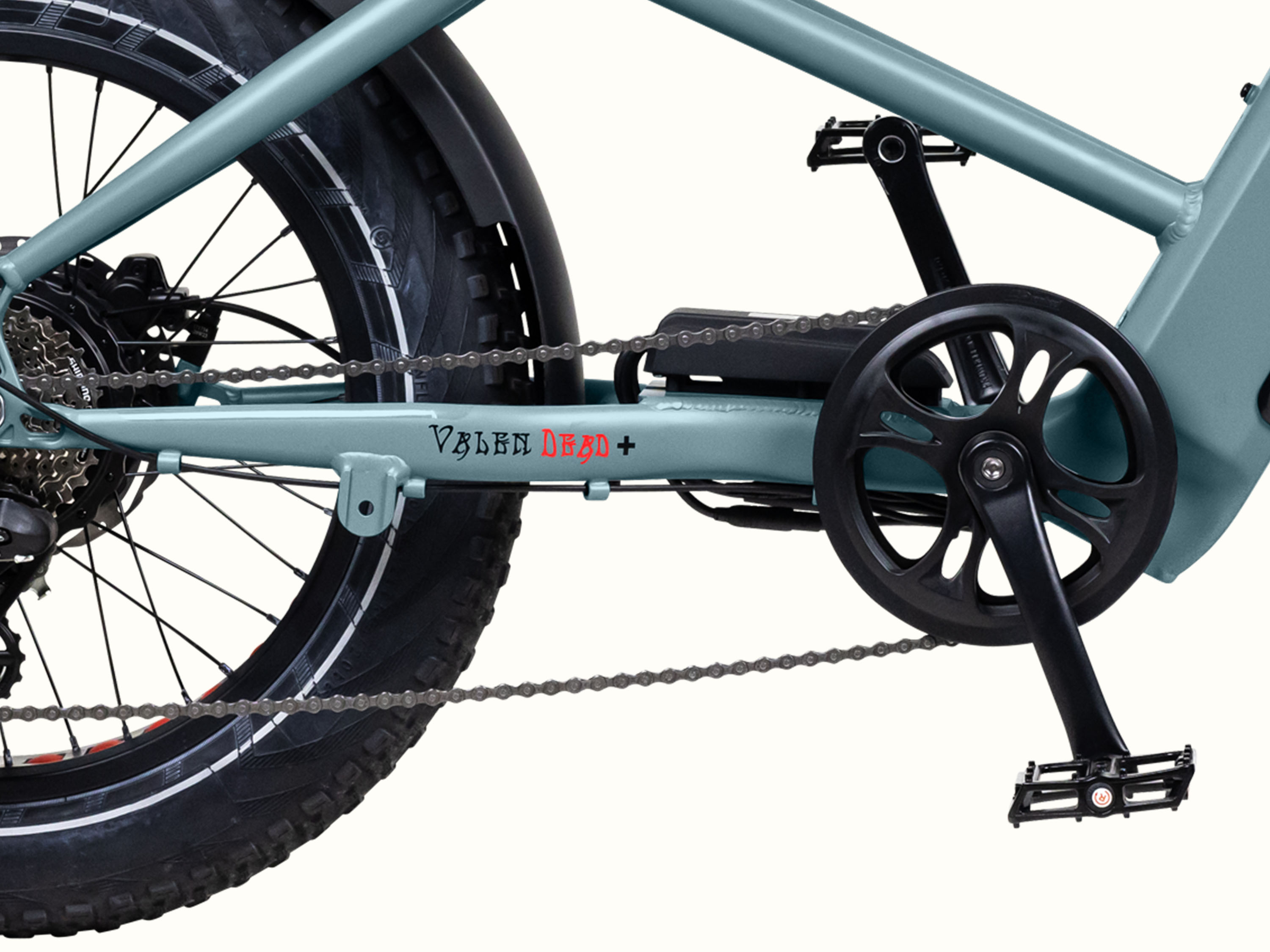 Close-up of the Grateful Dead Valen Rev+ Fat Tire EBike's drivetrain, featuring a light blue frame, black pedals, chain, and a fat rear tire. Text reads 