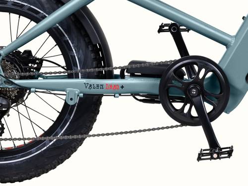 Close-up of the Grateful Dead Valen Rev+ Fat Tire EBike's drivetrain, featuring a light blue frame, black pedals, chain, and a fat rear tire. Text reads "Valen Dead+".