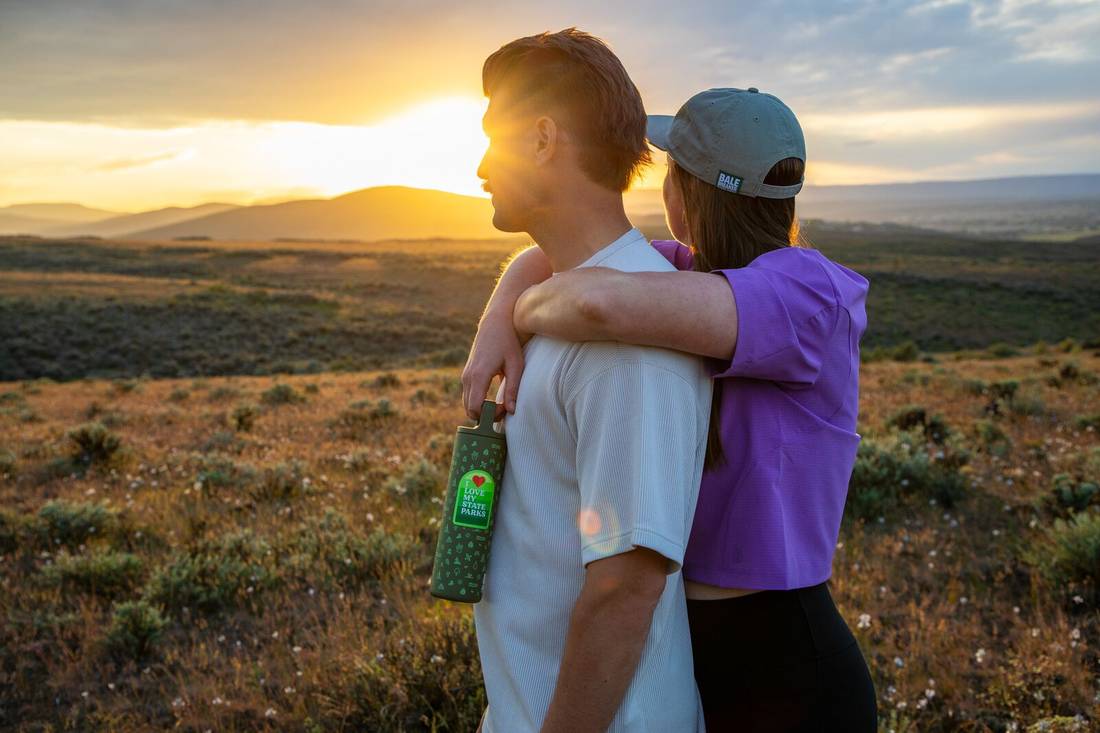 Person hugging another person from behind holding a MiiR x Bale Breaker Wide Mouth Bottle by handle and watching the sun set behind mountains