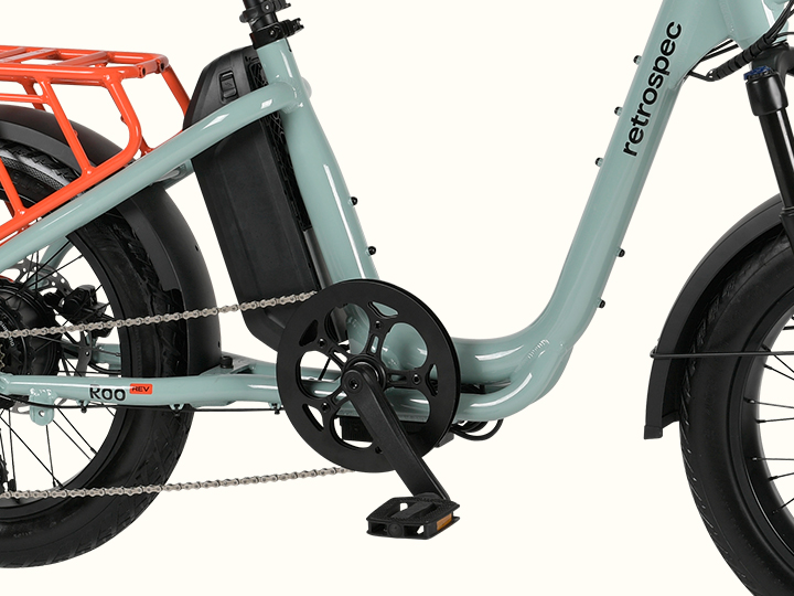 Close-up of a mint green electric bike with an orange rear rack, showcasing the pedal, chain, and battery compartment.