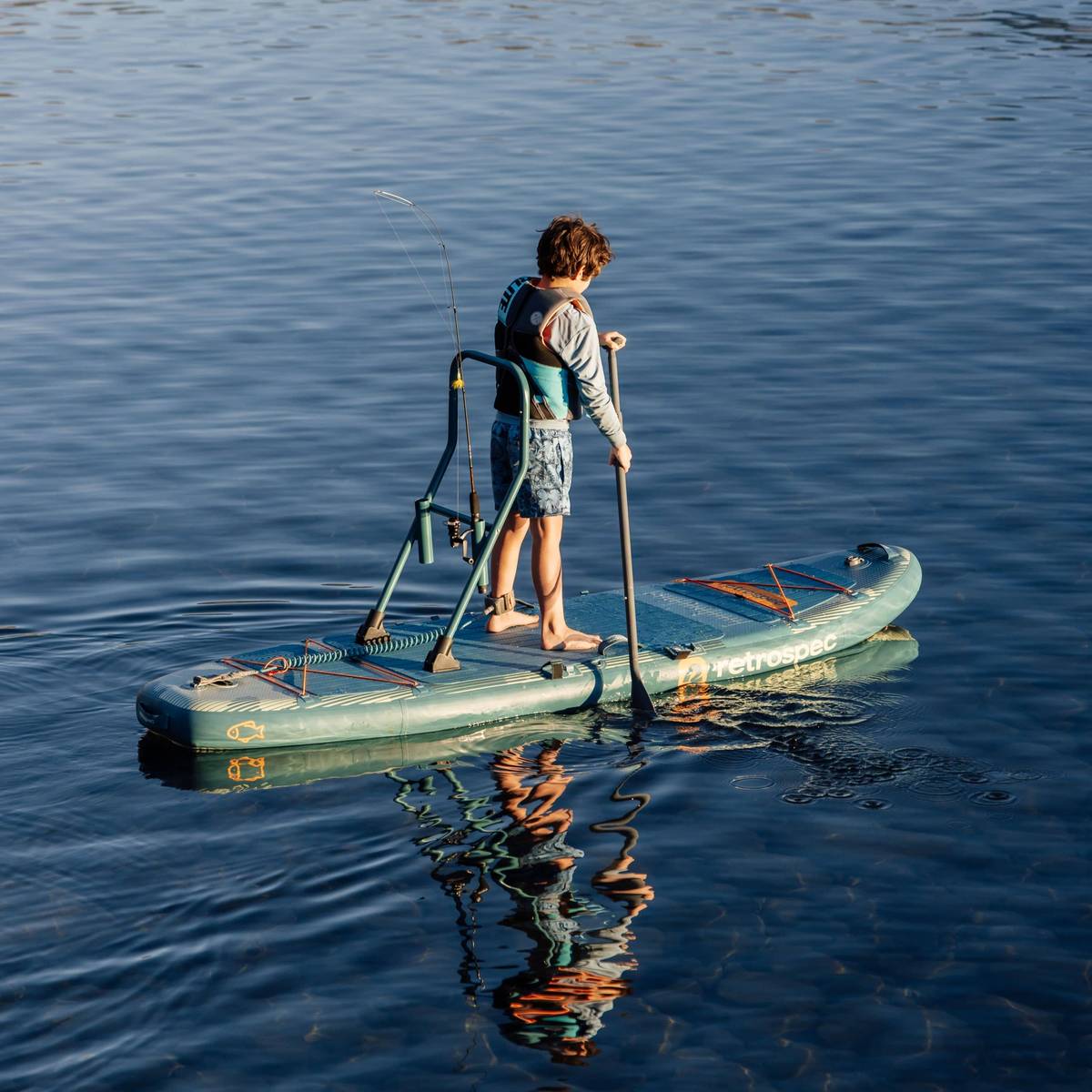 A child paddles while on a June Nano Kids Inflatable Stand Up Paddle Board Kayak Hybrid with 'retrospec' branding on a calm body of water.