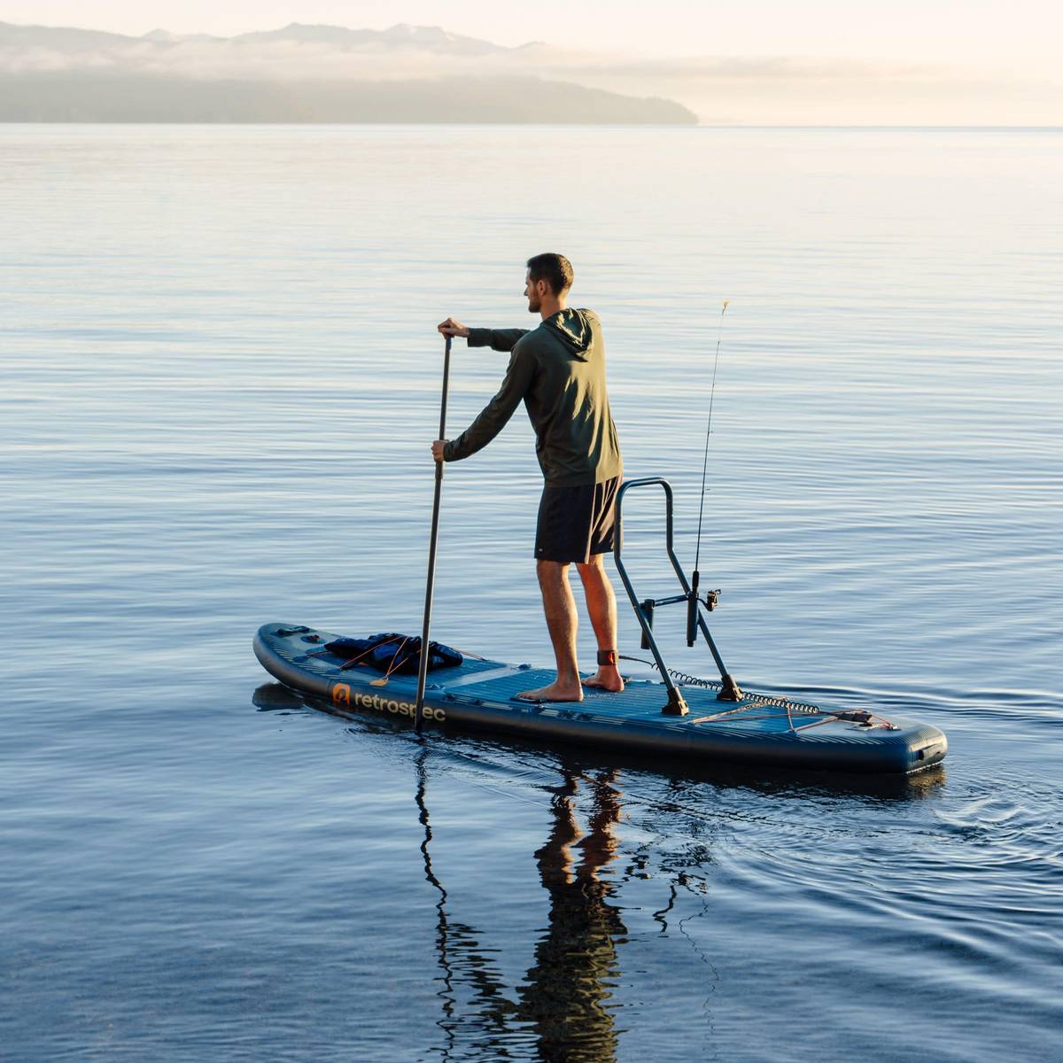 A person stands while paddling their June Inflatable Fishing Paddle Board Kayak Hybrid with the fishing rod holder attached to the board in calm water while the sun it setting.