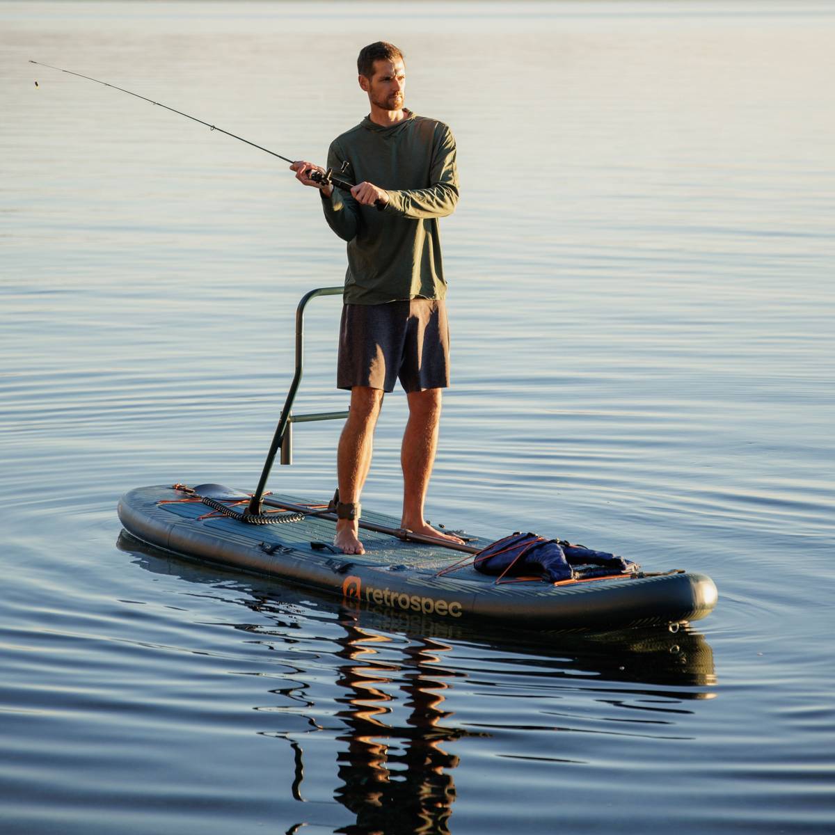 A person casts a fishing rode from a paddleboard in calm water, surrounded by a serene landscape at sunset.