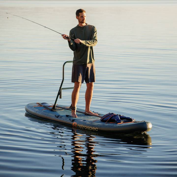 A person casts a fishing rode from a paddleboard in calm water, surrounded by a serene landscape at sunset.