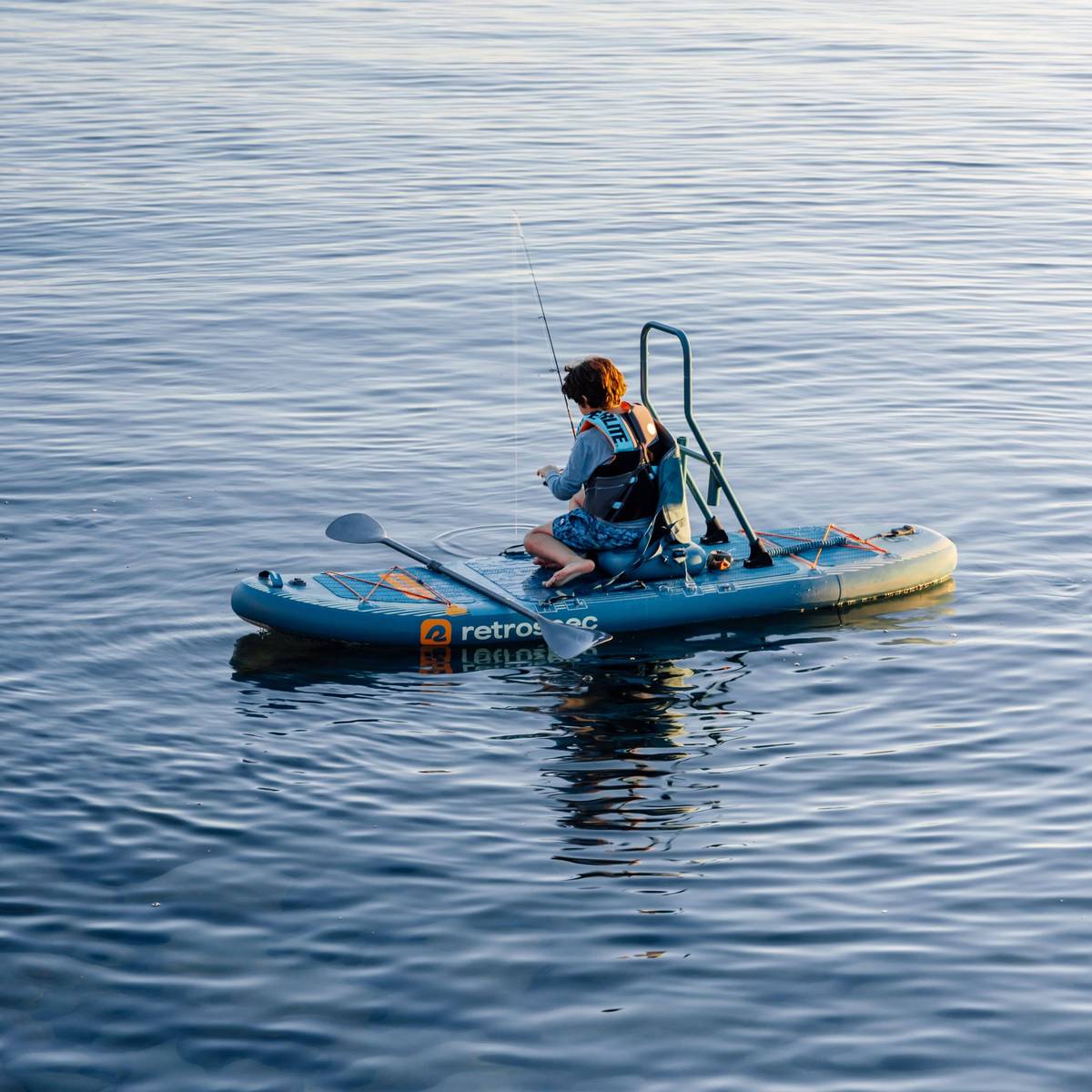 A kid sits on the blue June Nano Kids Inflatable Fishing Paddle Board Kayak Hybrid with fishing equipment on a calm body of water.