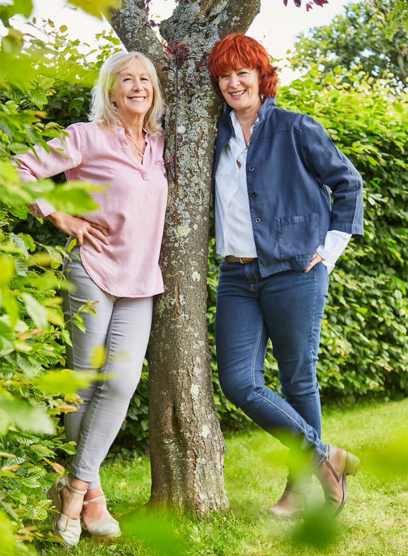 Susi Lennox and Sarah Brooks.  Founders of the YES YES Company standing in an English garden leaning against a tree looking happy and relaxed