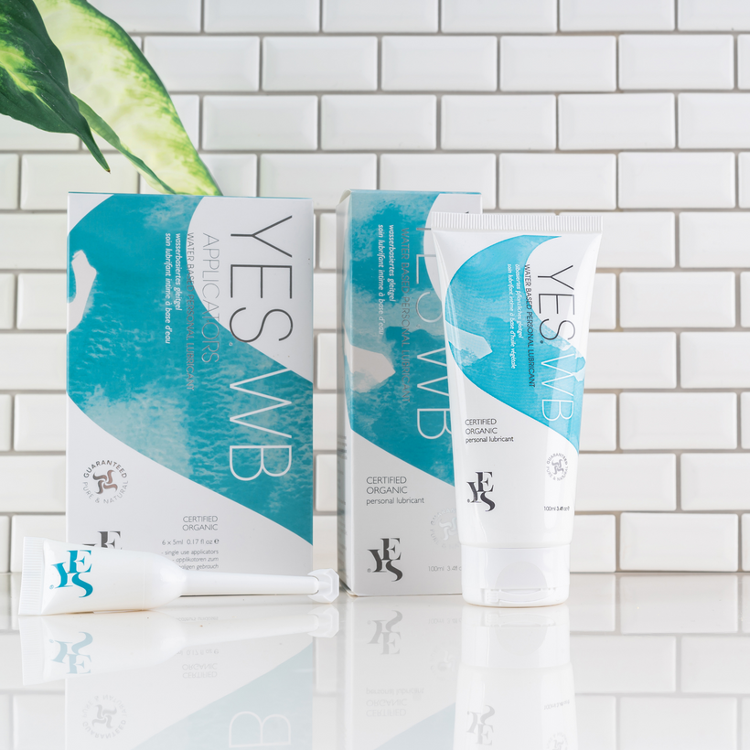 YES WB range stood against a white tiled wall and plant leaf in the background.  The range includes a tube of YES WB water-based lubricant, alongside a prefilled applicator of YES WB water-based lubricant.