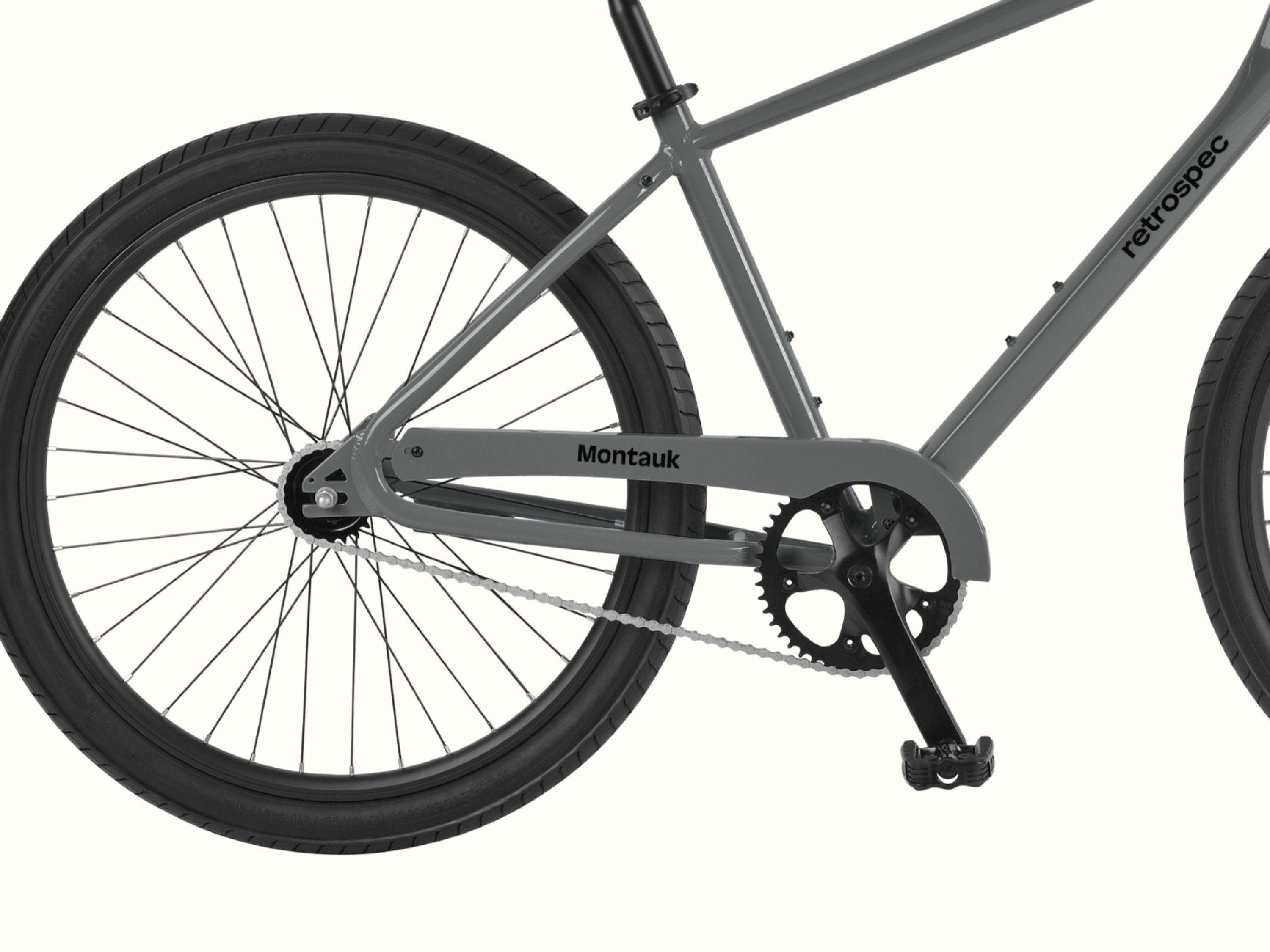 Close-up of the rear wheel, chain, and pedal area of a gray retrospec Montauk bicycle against a white background.