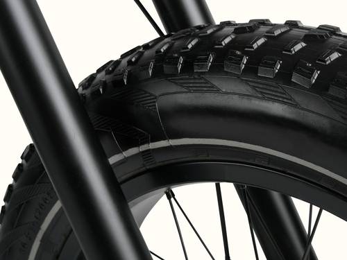 Close-up of a thick, rugged bicycle tire positioned between sturdy black fork legs, highlighting the tire's tread pattern and design.