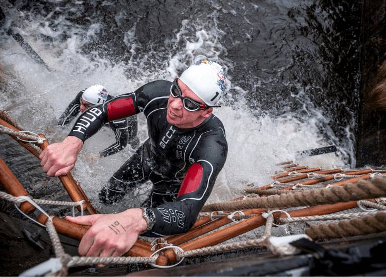 male swimmer wearing a HUUB wetsuit and swimming cap climbing a rope ladder to scale a canal wall