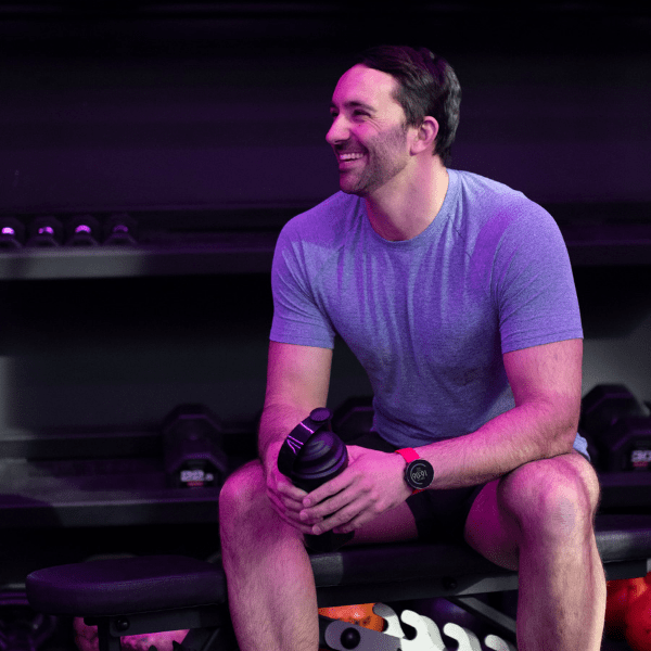Arron Collins-Thomas TONIQ founder and personal trainer sat in a purple lit gym on a weights bench holding a U Perform Shaker Bottle smiling to the camera