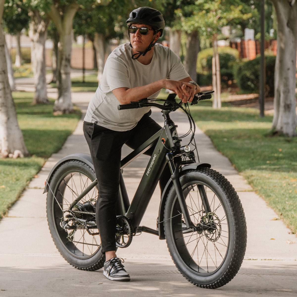A person in a gray t-shirt and black pants leans on a matte black Koa Rev 2 Fat Tire Electric Bike, surrounded by lush greenery along a paved path.