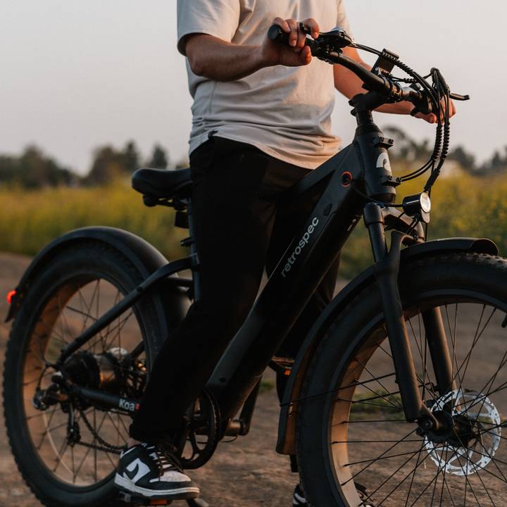 A person rides a matte black Koa Rev Fat Tire Electric Bike on a dusty trail, surrounded by greenery and a soft sunset glow.