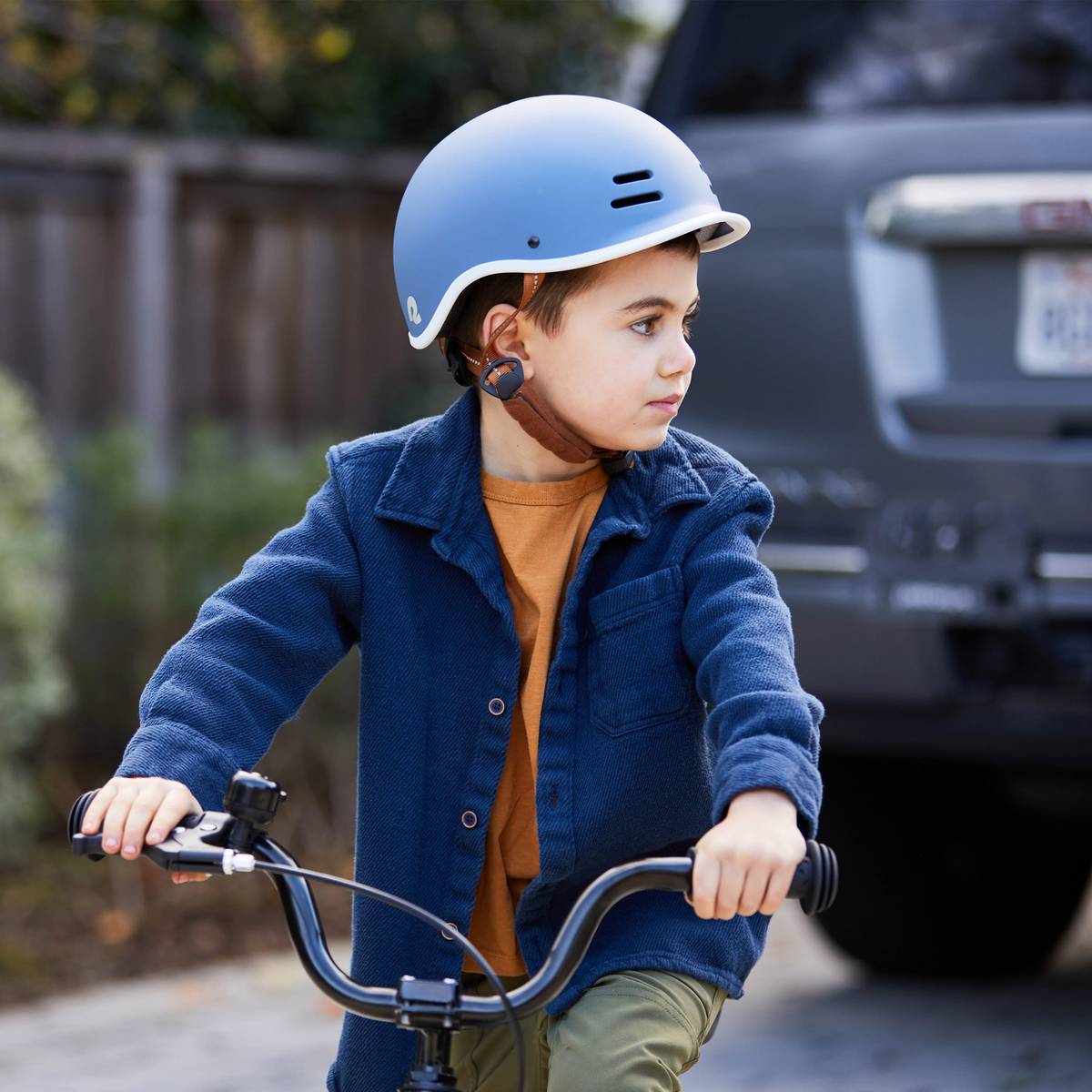 A child in a blue helmet rides a bicycle, wearing a blue jacket over an orange shirt, with earbuds in a residential area.