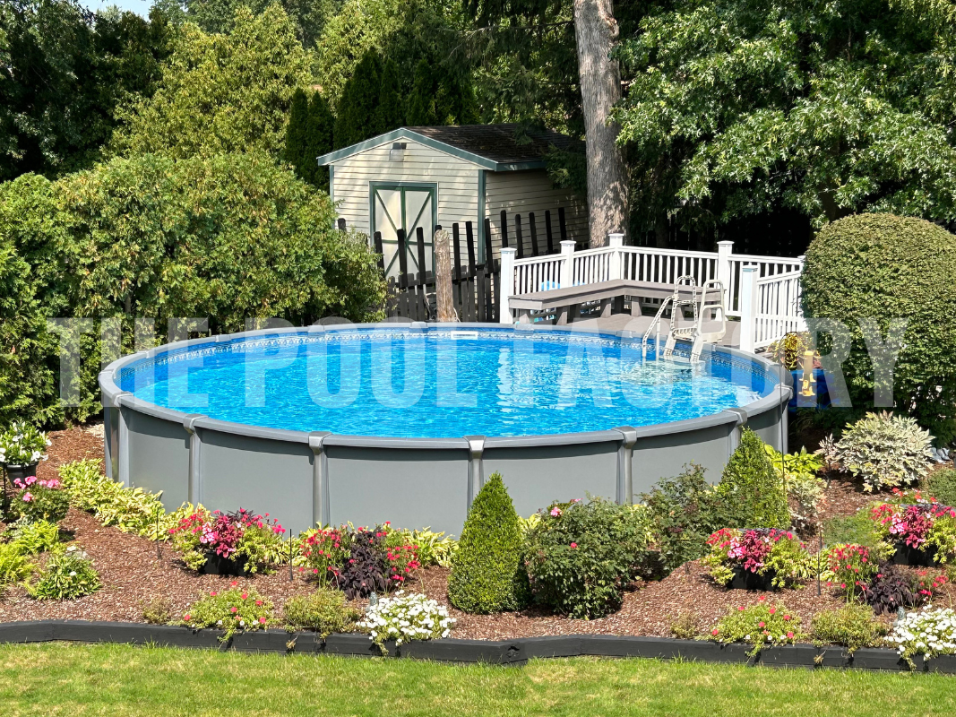 Round above ground pool with partial deck, colorful flowers, shrubs, and a lush forest landscape