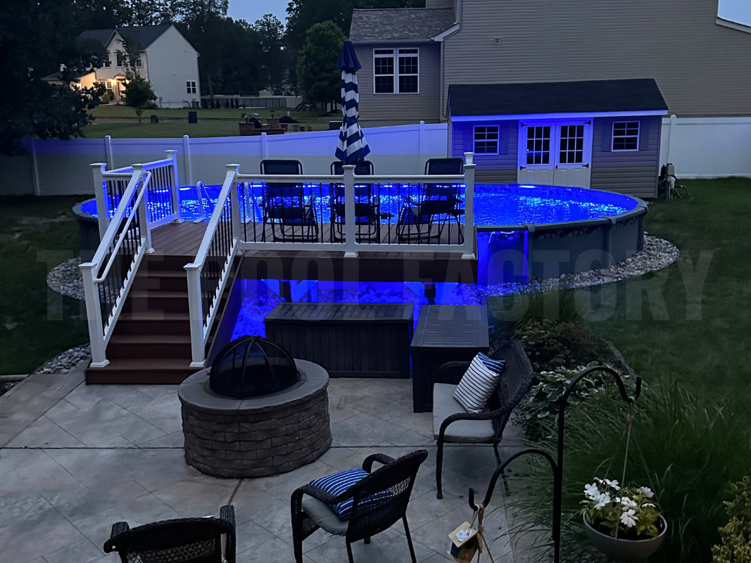 Oval semi-inground saltwater pool with fire pit, partial deck, and lights at night