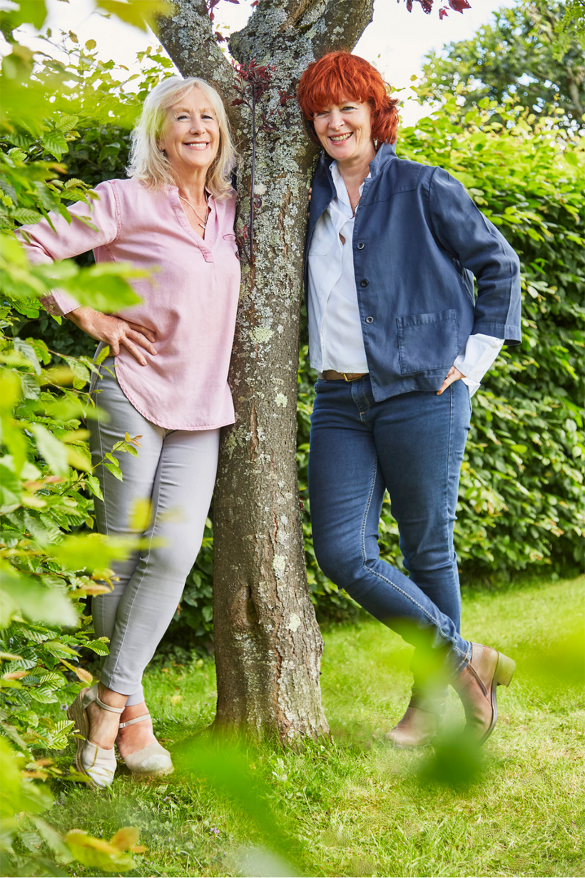 Susi Lennox and Sarah Brooks.  Founders of the YES YES Company standing in an English garden leaning against a tree looking happy and relaxed