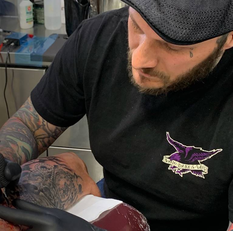 Tattoo artist in a cap works on a detailed arm tattoo.