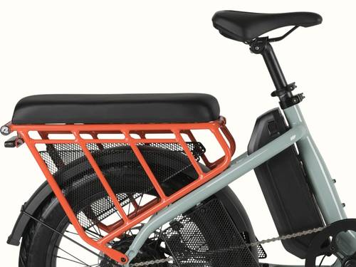 Close-up of the retrospec Roo Rev electric cargo bike showing the black passenger seat pad installed on the orange rear rack with wheel guard set and integrated battery.
