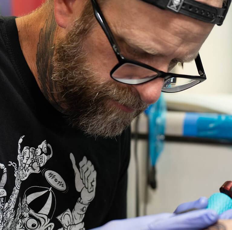 Tattoo artist wearing glasses and gloves, focusing intently on work.