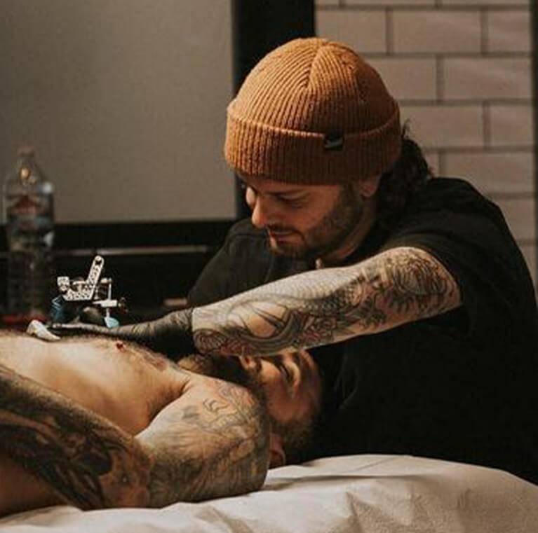 Tattoo artist with tattoos, wearing a brown beanie, working on a man's tattoo.