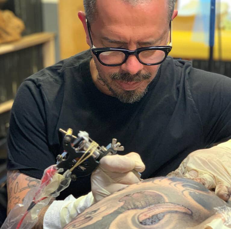 Tattoo artist with glasses focusing on tattooing a client's arm.