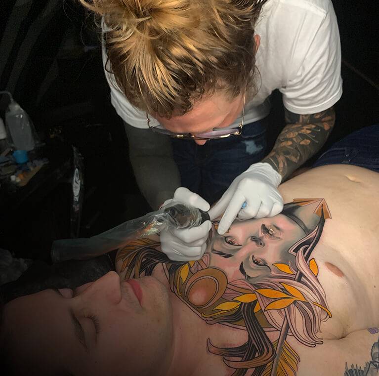 Tattoo artist working on a detailed chest design of a woman's face with ornate elements.