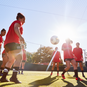 Female footballer wearing red shirt preparing to strike a football that is coming back down towards the ground from the air