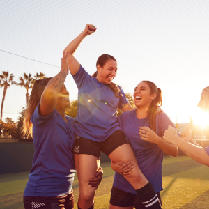 Group of female footballers wearing blue shirts and black shorts celebrating a goal by lifting the scorer in the air on their shoulders