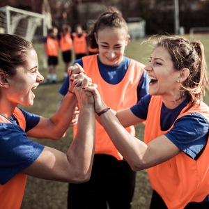 Group of female footballers wearing blue shirts and an orange vest celebrating a goal by high fiving each other
