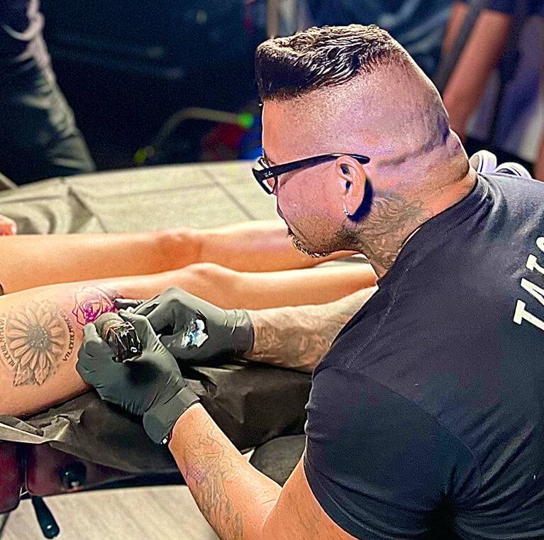 Tattoo artist with glasses and gloves, adding colour to a rose tattoo on a person's arm.