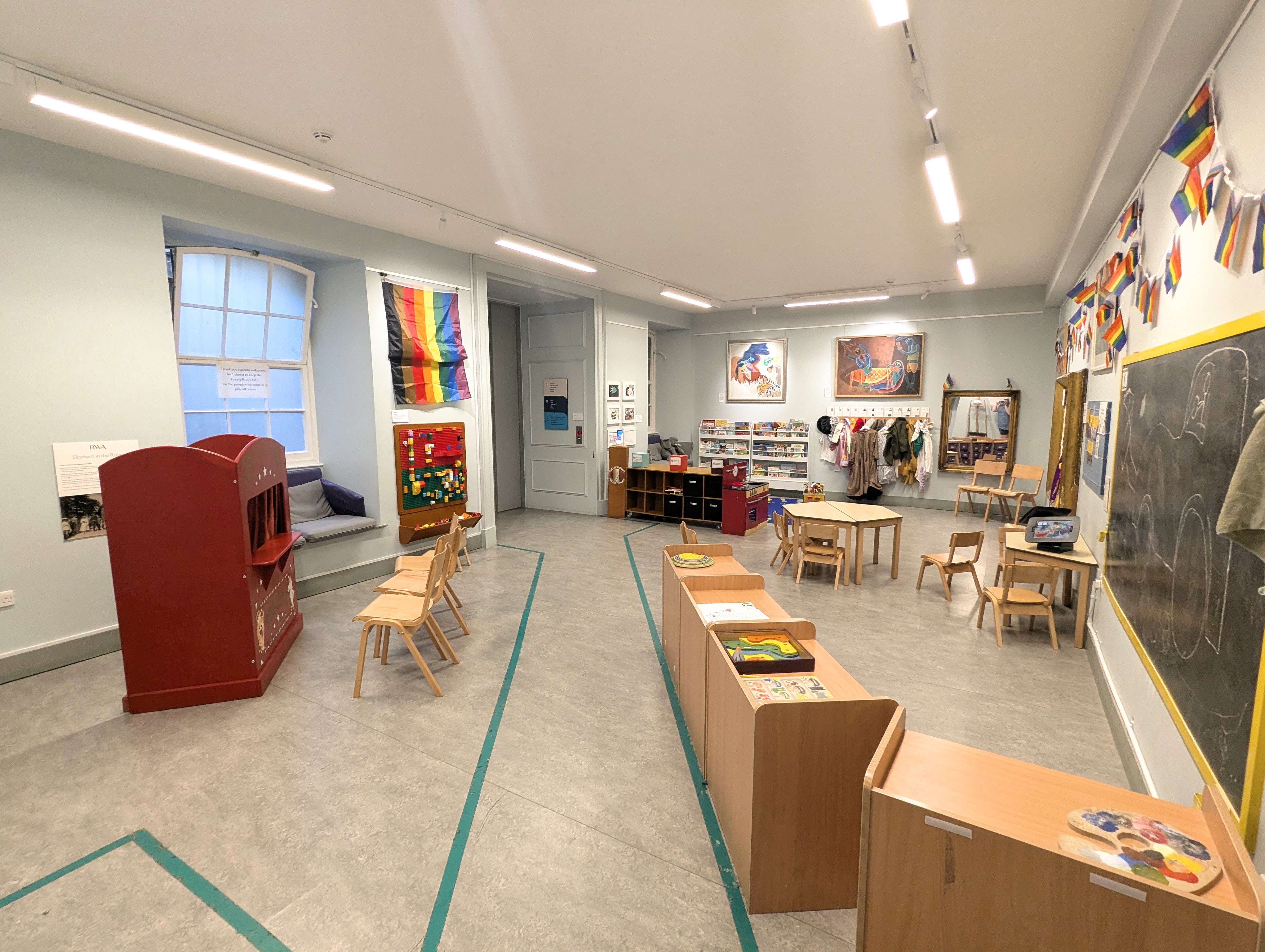 A large room with artwork on the walls, tables and chairs and a blackboard to the side of the space 