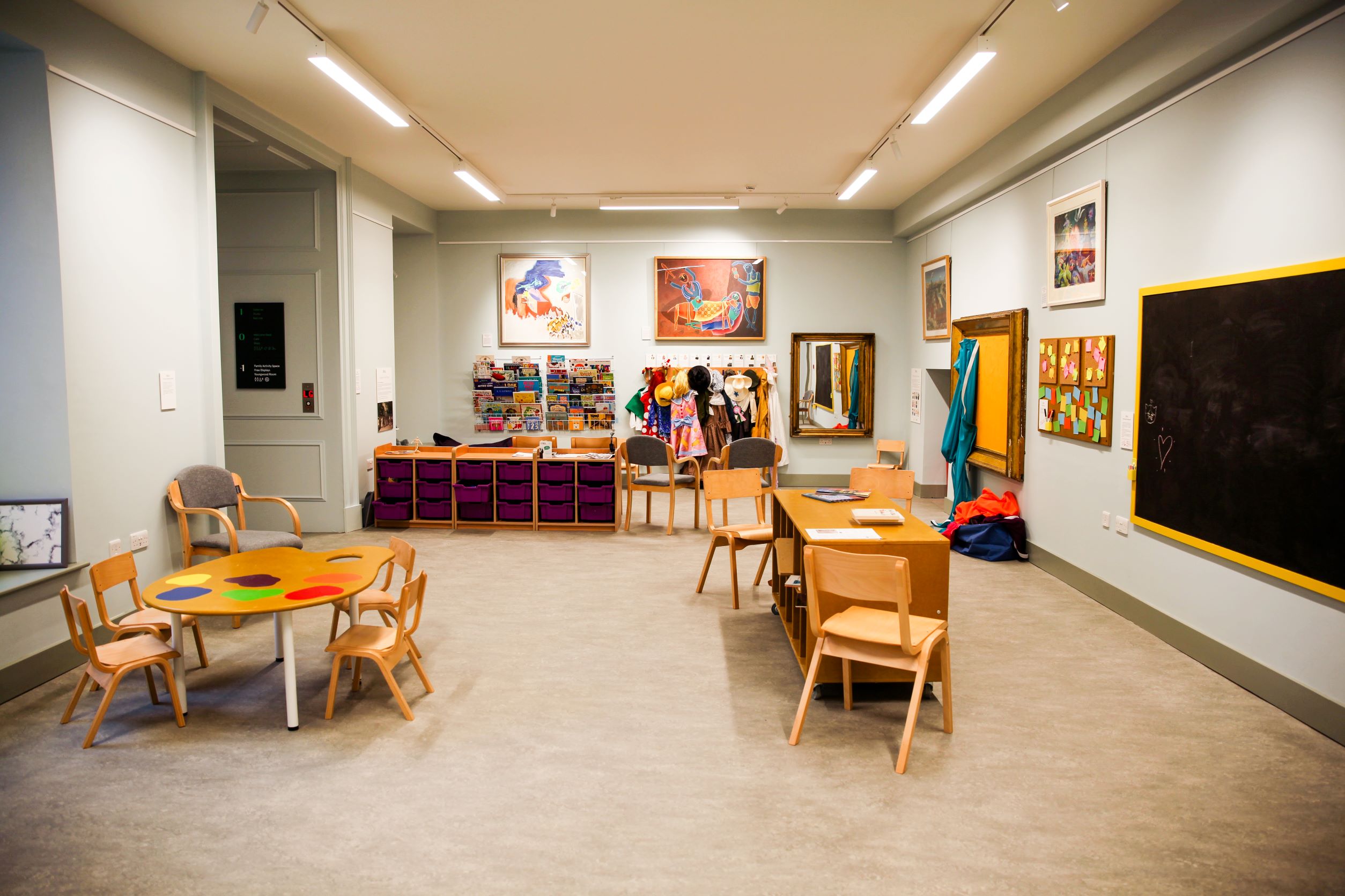 A large room with artwork on the walls, tables and chairs and a blackboard to the side of the space 