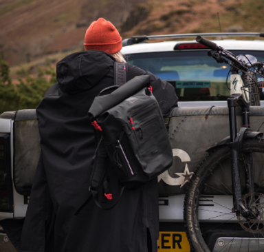 Woman wearing Red Equipment Voyager Beanie in Orange, Long Sleeve Pro Change Robe EVO in Stealth Black and carrying the Adventure Waterproof Backpack in Obsidian Black unloading a bike from the back of a pickup truck.