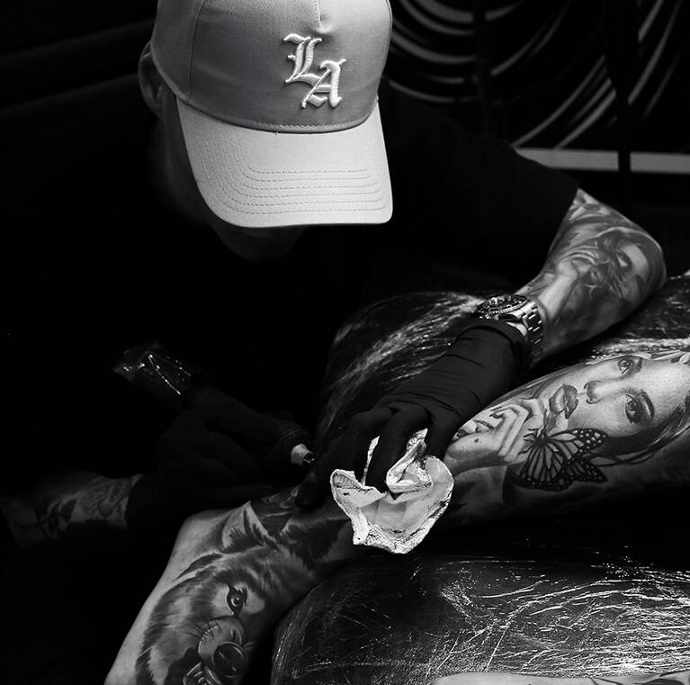 Tattoo artist working on a detailed leg tattoo, wearing a cap and gloves.