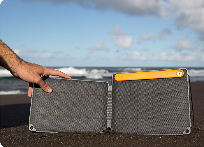 A hand holds a portable solar panel on a beach, with waves and a cloudy sky in the background.